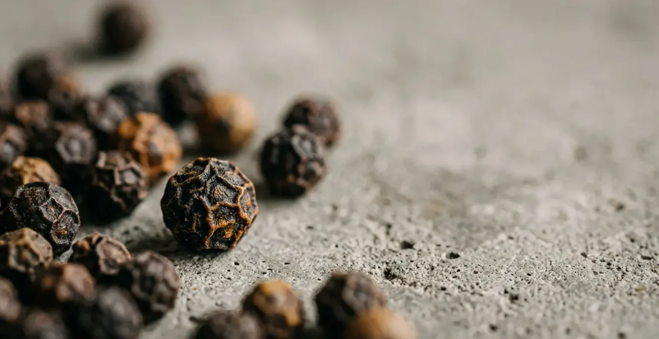Gros plan sur des grains de poivre noir artisanaux éparpillés, montrant leurs textures ridées et leur couleur intense